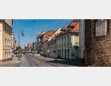 Straßenszene einer Altstadt mit gepflastertem Straßenbelag, Straßenbahnschienen in der Mitte und historischen Gebäuden mit roten Ziegeldächern auf beiden Seiten. Rechts im Vordergrund steht ein altes Backsteintor mit einer Gedenktafel, während im Hintergrund Cafés und Schilder zu sehen sind.