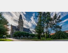 Historisches Gebäude mit einem hohen Uhrturm, umgeben von gepflegtem Grün und großen Bäumen, unter einem Himmel mit weißen Wolken; im Vordergrund führt ein Weg durch eine Parkanlage.