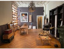 Gemütlicher Raum mit Holzdielenboden, einem Holztisch mit Stühlen und stilvollen Regalen an den Wänden. Im Vordergrund steht ein Sessel mit Fell und ein kleiner Couchtisch; die Wand zeigt moderne Kunstwerke und Beleuchtung, während der Raum hohe Decken mit dekorativen Elementen hat.