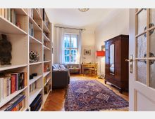 Wohnzimmer mit gemütlicher Einrichtung, bestehend aus einem grauen Sofa, einem Bücherregal, einem Holztisch mit Stuhl und einem großen Teppich mit orientalischem Muster auf einem Parkettboden. Ein Fenster mit weißen Vorhängen und ein dunkler Holzschrank prägen den Raum, der von einer Deckenlampe und einer roten Stehlampe beleuchtet wird.