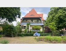 Zweistöckiges Wohnhaus mit rotem Satteldach und einem großen Balkon, der von einem blauen Holzgeländer umgeben ist. Das Haus ist von einem Garten mit üppiger Vegetation umgeben, im Vordergrund befindet sich ein kleiner gepflasterter Platz mit einem abgedeckten Motorrad.