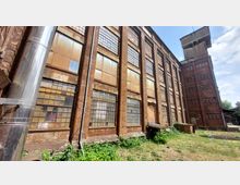 Backstein-Industrielager mit großen rechteckigen Fenstern, teils intakt und teils mit Patina versehen, sowie einer angebauten hohen, schlanken Turmstruktur. Der Hof im Vordergrund ist überwuchert von Gras und Pflanzen, Metallrohre verlaufen entlang der Fassade, und der Himmel ist teils bewölkt.