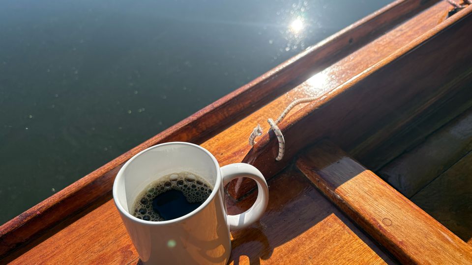 Holzboot auf ruhigem Wasser, mit glänzendem Bootsrand und Sonnenreflexionen auf der Wasseroberfläche. Im Vordergrund steht eine weiße Kaffeetasse auf einer hölzernen Sitzbank im Boot.