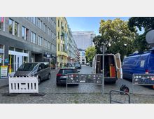 Straßenszene in einer städtischen Umgebung mit Kopfsteinpflaster; links moderne Gebäude mit Geschäften, rechts parkende Fahrzeuge, darunter ein offener weißer Lieferwagen und ein blauer Transporter. Im Hintergrund ist das Logo der „Charité“ auf einem großen Gebäude sichtbar, umgeben von Bäumen und kleineren Gebäuden.