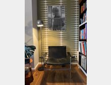 Ecke eines Wohnzimmers mit einem schwarzen Ledersessel auf einem Holzfußboden, daneben eine Stehlampe mit metallischem Lampenschirm. Der Hintergrund besteht aus einer gemusterten Tapete in Olivgrün und Weiß, darauf hängt ein Schwarz-Weiß-Bild. Rechts sind Bücherregale mit Büchern zu sehen.
