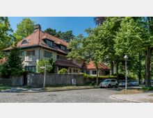 Straßenszene in einem Wohnviertel mit einem großen, historischen Haus im Stil der frühen Moderne, das durch ein rotes Ziegeldach und weiße Fassaden gekennzeichnet ist. Umgeben von Bäumen, sind am Straßenrand geparkte Autos und eine Straßenlaterne sichtbar.