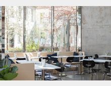 Ein modernes Café mit hellen Holzmöbeln und schwarzen Stühlen, großen Fenstern, die Blick auf einen begrünten Außenbereich und Gebäude bieten. Zusätzlich sorgen die Betonwände und Tageslicht für ein minimalistisches Ambiente.
