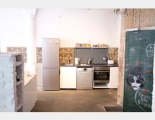 Kleine Küche in einem industriellen Stil mit weißen Wänden, grauem Boden und Mosaikfliesen an der Rückwand. Zu sehen sind ein silberner Kühlschrank, Spüle, Geschirrspüler, Backofen und offene Regale; an der rechten Wand ein schwarzes Brett mit Kreidezeichnungen und Graffiti.