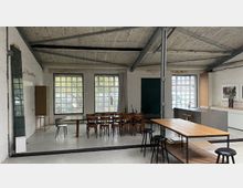 Großer, offener Raum im Industrie-Stil mit Sichtbetondecke, Stahlträgern und großen Glasbausteinfenstern, die viel Tageslicht hereinlassen. Im Inneren stehen mehrere Tische mit Stühlen und Hockern sowie eine lange Arbeitsfläche entlang der rechten Wand.