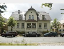 Zweistöckiges Wohnhaus mit hell beigem Putz, symmetrischer Fassade, Satteldach und Gaubenfenstern. Vor dem Gebäude stehen mehrere geparkte Autos, und es ist von einem Zaun sowie kleinen Bäumen umgeben.