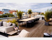 Dachterrasse mit Holzboden, einem langen Esstisch und schwarzen Stühlen, umgeben von Pflanzkübeln mit Bambus und kleinen Bäumen; im Hintergrund eine Outdoor-Küche mit Grill und Blick auf umliegende Gebäude einer Stadt.