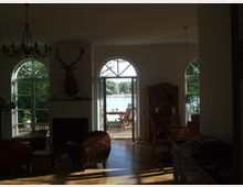 Wohnzimmer mit großen, weißen Rundbogenfenstern, durch die Blick auf einen Holzbalkon und einen See mit Ufervegetation sichtbar ist. Im Raum stehen ein Ledersessel, ein Kamin mit Hirschgeweih an der Wand, ein hölzerner Schrank und ein Kronleuchter an der Decke.