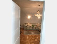 Wohnzimmer mit glänzendem Parkettboden, einer hellen Eckcouch mit dekorativen Kissen, einem Glastisch mit Pflanzen darauf und einem großen Kronleuchter an der Decke. Die Wände sind hell gestrichen, und eine offene weiße Tür gibt den Blick in den Raum frei.