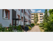 Mehrfamilienhäuser in einer Wohnsiedlung mit gepflastertem Gehweg dazwischen, umgeben von gepflegten Grünflächen, Bäumen und Sträuchern; moderne Balkone und bunte Fensterrahmen sind sichtbar.