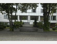 Modernes Gebäude mit einer Fassade in weißer Plattenoptik, unterstützt von weißen Säulen im Eingangsbereich. Vor dem Gebäude befindet sich ein gepflasterter Weg mit kleinen bepflanzten Beeten und großen Bäumen, die teilweise mit Efeu bewachsen sind.