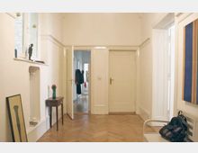 Heller Flur mit Holzboden in Fischgrätmuster, weißen Wänden und mehreren Türen, die zu angrenzenden Räumen führen. Links steht ein kleiner Tisch mit einer Vase, ein hochkant angelehnter Bilderrahmen und eine schmale Fensterbank mit Dekorationen; rechts befindet sich eine bunte Wanddekoration und eine Sitzbank mit einem Rucksack darauf.