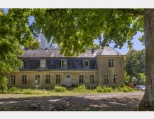 Zweistöckiges, verwittertes Gebäude mit schrägem Dach und mehreren Fenstern, umgeben von dichtem Laubbaumwald, mit einem unbefestigten Weg im Vordergrund.