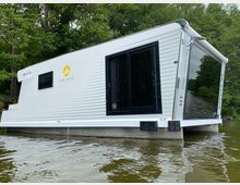 Modernes Hausboot auf einem ruhigen Gewässer, umgeben von dichtem Wald. Das Boot hat eine weiße Fassade mit schwarzen Fenstern und trägt die Aufschrift „The Kiss“.