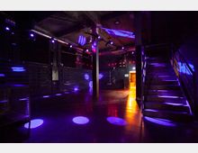 Ein leerer, dunkler Clubraum mit Betonwänden und -decke, beleuchtet durch bunte Lichtspots in Lila- und Blautönen. Rechts sind eine Metalltreppe und ein beleuchteter Durchgang zu sehen, während die Decke mit Lüftungsrohren und Beleuchtungssystemen ausgestattet ist.