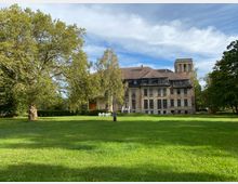 Eine historische Backsteinvilla mit großen Fenstern und einem Turm steht inmitten einer weitläufigen, grünen Wiese, umgeben von großen Bäumen unter blauem, teils bewölktem Himmel.
