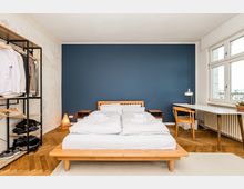 Helles Schlafzimmer mit Doppelbett und Holzrahmen vor einer dunkelblauen Akzentwand, flankiert von zwei Nachttischen. Rechts stehen ein Schreibtisch mit Stuhl vor großen Fenstern und Heizkörper, links ein offenes Kleiderregal; der Boden ist im Fischgrätparkett verlegt.