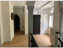 Flur mit heller Wandgestaltung, teppichartigem Bodenbelag und gewölbten Durchgängen. Auf der rechten Seite befinden sich weiße Türen mit Sprossenfenstern und ein Heizkörper unter einer halbkreisförmigen Fensteröffnung.