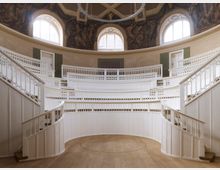 Rundes, historisches Auditorium mit weißen, hölzernen Sitzreihen und Geländern, umgeben von Wandmalereien und großen, bogenförmigen Fenstern, die Tageslicht hereinlassen. Der Boden besteht aus hellem Holz, und die Architektur ist symmetrisch und detailreich gestaltet.