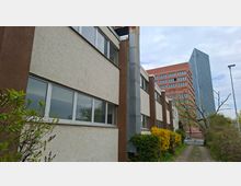 Schmaler Gehweg entlang eines flachen Büro- oder Verwaltungsgebäudes mit heller Putzfassade, langen Fensterbändern und einem senkrechten Metallschacht. Im Hintergrund stehen ein rotes Hochhaus und ein schlanker gläserner Büroturm, umgeben von Sträuchern und Bäumen.