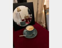 Eine grüne Kaffeetasse mit passendem Unterteller und Teelöffel steht auf einem dunkelroten Tischtuch vor einem runden Spiegel, der das Getränk und den Hintergrund mit einer Stehlampe und weiteren Objekten reflektiert. Rechts im Bild befindet sich ein Bücherstapel auf einem Holzständer.