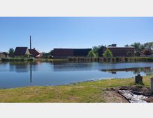 Ein Dorfteich mit ruhiger Wasseroberfläche, im Hintergrund mehrere ländliche Gebäude mit roten Ziegeldächern und ein hoher Schornstein. Grüne Vegetation und Schilf säumen das Ufer.