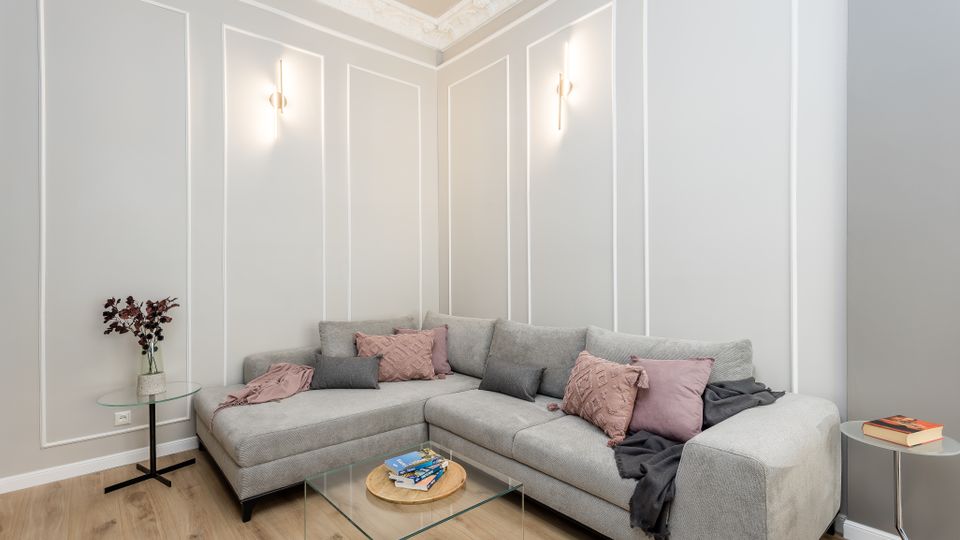 Modernes Wohnzimmer mit hellgrauer Eckcouch, Glas-Couchtisch und kleinen Beistelltischen auf Holzfußboden. Hohe Wände mit weißen Zierleisten und Stuckleiste an der Decke sowie zwei schmale Wandlampen prägen den Raum.