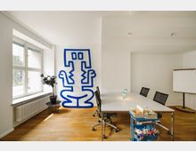 Heller Konferenzraum mit Holzboden, einem großen weißen Tisch und schwarzen Stühlen. An der Wand ein markantes blaues Wandkunstwerk, links ein Fenster mit Heizkörper sowie ein Beistelltisch mit einer Pflanze; rechts ein Flipchart und ein Rollwagen mit Büromaterialien.