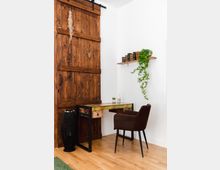 Ein kleiner Arbeitsbereich mit einem rustikalen Holztisch und einem braunen Polsterstuhl auf einem hellen Holzboden. Links befindet sich eine Holzschiebetür, rechts hängt ein Regal mit Büchern und einer rankenden Pflanze.