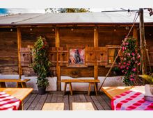 Rustikale Holzfassade einer Hütte mit kleinen Fenstern und Fensterläden mit herzförmigen Ausschnitten sowie einer Lichterkette unter dem Dach. Davor stehen Holzbänke und Tische auf einer Terrasse, flankiert von großen Topfpflanzen und blühenden Kletterpflanzen.