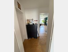 Schmaler Flur mit weißen Wänden und Holzdielenboden, links eine schwarze Schuhkommode mit Pinnwand und kleiner Dekoration. Durch eine offene Tür blickt man in ein helles Wohnzimmer mit großem Fenster, Teppich und einer Zimmerpflanze.