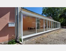 Ein Gebäude mit rosa Fassade und einem langen, modernen Wintergarten aus weißen Rahmen und großen Glasscheiben. Vor der Glasfront stehen Tische und Stühle, das Umfeld ist mit Kopfsteinpflaster und Grünflächen gestaltet.