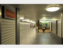 Unterirdischer Durchgang mit Fliesenwänden und Deckenlampen; links ein Schild mit der Aufschrift „Geldautomat“, rechts ein Blumenladen mit der Aufschrift „Blumen & Co“ sowie Auslagen mit Blumenarrangements.