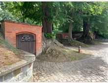 Ein kleiner, hügeliger Bereich mit Bäumen, auf dem sich mehrere in den Boden eingelassene Weinkeller mit roten Ziegelbögen und dunklen Holztüren befinden. Im Vordergrund ist ein gepflasterter Weg und eine niedrige Mauer aus Natursteinen sichtbar.