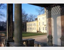 Gelbes klassizistisches Gebäude mit symmetrischen Fenstern, umgeben von einer Rasenfläche und kahlen Bäumen im Hintergrund. Im Vordergrund stehen Säulen und ein Teil einer steinernen Balustrade.