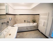 Helles Badezimmer mit grauen Steinfliesen, eingelassener Badewanne an der Rückwand und großem Spiegel über einem breiten Waschbecken. Rechts steht ein weißer Handtuchheizkörper; weiße Decke und schwarze Fliesenbordüre rahmen den Raum.