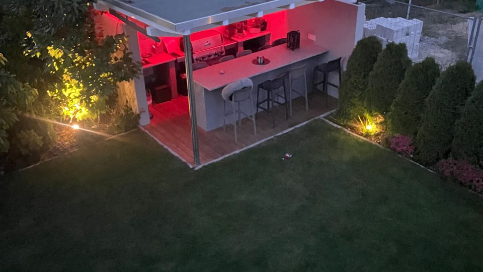Moderner Garten mit einer kleinen offenen Gartenküche unter einem Flachdach, beleuchtet durch rotes und warmweißes Licht. Im Vordergrund liegt eine Rasenfläche, umgeben von hohen Bäumen und Hecken.