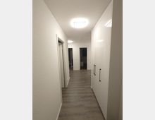 Schmaler, gut beleuchteter Flur in einer Wohnung mit einem modernen Vinylboden und weißen Wänden. Rechts befindet sich ein Einbauschrank mit glänzenden weißen Türen, links führen mehrere Türen in angrenzende Räume.