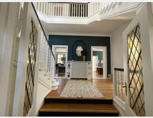 Eingangsbereich eines Hauses mit weiß lackierten Holzgeländern und Holztreppe. Glasschiebetüren mit Rautenmuster flankieren den Raum, der eine dunkelgrüne Akzentwand, eine Kommode mit ovalem Spiegel und einen hellen Teppich über Parkettboden zeigt. Im Hintergrund sind weitere Räume mit klassischer Einrichtung sichtbar.