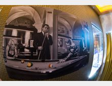 Eine Innenaufnahme mit einer großflächigen Wandmalerei in Schwarz-Weiß, die eine Szene mit Personen an einem Billardtisch darstellt. Neben der Malerei befinden sich ein beleuchtetes Regal und gelbe Vorhänge.