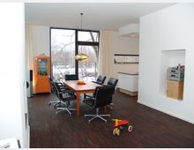 Esszimmer in einem modernen Wohnraum mit einem Holztisch, umgeben von schwarzen Lederstühlen auf einem dunklen Holzboden. Große Fenster bieten Blick auf einen schneebedeckten Garten, während eine orangefarbene Vitrine und eine offene Küche den Raum ergänzen.