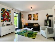 Wohnzimmer mit modernem Design, hellen Wänden und großen farbigen Gemälden. Eine schwarze Ledercouch, ein weißer Sessel, ein Teppich mit Blattmuster, ein Holzofen sowie große Fenster, die Tageslicht hereinlassen, prägen den Raum.