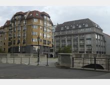 Zwei mehrstöckige Gebäude in urbaner Umgebung, darunter ein gelbes Wohnhaus mit roten Ziegeldächern und großen Fenstern sowie ein graues Gebäude mit neoklassizistischen Details. Im Vordergrund verläuft eine Brücke mit Geländern und Laternen.