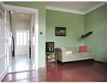 Wohnzimmer mit hellgrünen Wänden, Holzboden in dunklem Rot, einem beigen Sofa mit roten und weißen Kissen, einer Wandlampe und einem kleinen Beistelltisch mit einem Retro-Radio. Im Hintergrund ist eine geöffnete Tür zu einem Flur mit Tageslicht durch eine Glastür sichtbar.