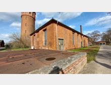 Backsteingebäude mit hohen Fenstern und einer Holztür, daneben ein runder Wasserturm aus Backstein. Im Vordergrund eine rostige Metallfläche und ein gemauerter Vorsprung, im Hintergrund eine asphaltierte Straße und Bäume.