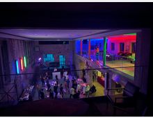 Industrieller Veranstaltungsraum mit sichtbaren Backsteinwänden, einer Tanzfläche im Erdgeschoss und einer Galerie im Obergeschoss, beleuchtet mit bunten Neonlichtern. Menschen tanzen unten, während oben Sitzgelegenheiten und farbige Beleuchtung die moderne Atmosphäre betonen.